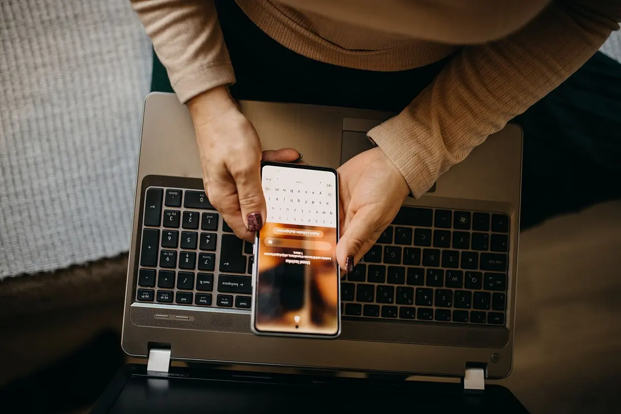 Manos revisando un teléfono sobre un notebook.